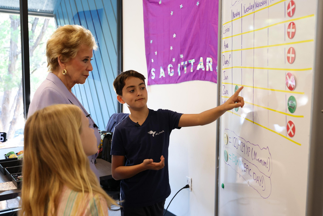 A boy points to a whiteboard chart with names and symbols while an older woman and a girl watch attentively in a classroom.